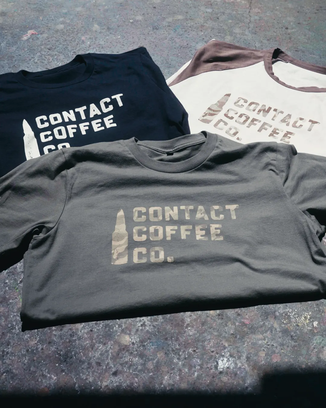 Three Contact Coffee tees laid out on concrete